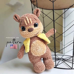 May include: A crocheted brown and tan plush toy with a yellow vest. The toy has a friendly face with blue eyes and a brown nose. The toy is standing on a white surface next to a black wire basket.