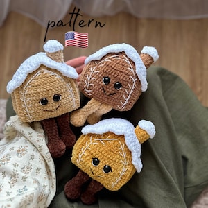 Könnte beinhalten: Drei gehäkelte Lebkuchenhaus-Spielzeuge mit weißem Zuckerguss und lächelnden Gesichtern. Die Häuser sind braun, beige und gelb. Das Bild enthält den Text "pattern" und eine amerikanische Flagge.