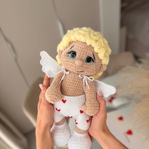 Puede incluir: Muñeca de ganchillo hecha a mano de Cupido, con tono de piel claro, cabello rizado amarillo y ojos azules. La muñeca lleva pantalones cortos blancos con corazones rojos y calcetines blancos. Tiene alas blancas.