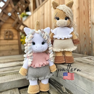 Pode incluir: Duas bonecas de unicórnio de crochê feitas à mão. Uma é branca com um top rosa e uma saia cinza, a outra é bege com um top branco e uma saia bege. Ambas têm cabelo encaracolado e estão em pé sobre uma superfície de madeira. A palavra "pattern" está escrita na imagem com a bandeira americana.