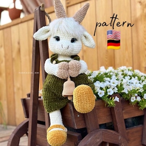 Puede incluir: Una muñeca de cabra de crochet que lleva un atuendo verde y marrón con una campana. La muñeca está sentada en un carro de madera con flores blancas en el fondo. El texto "pattern" está escrito en la esquina superior derecha de la imagen. También se muestran las banderas de Estados Unidos y Alemania.