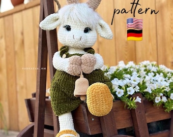 Op de afbeelding: Een gehaakte geitenpop die een groene en bruine outfit draagt met een bel. De pop zit op een houten kar met witte bloemen op de achtergrond. De tekst "pattern" staat in de rechterbovenhoek van de afbeelding. De Amerikaanse en Duitse vlaggen zijn ook te zien.