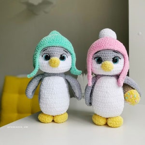Può includere: Due giocattoli di pinguino all'uncinetto, uno con un cappello verde chiaro e l'altro con un cappello rosa. Entrambi i pinguini hanno un corpo bianco, una testa grigia e un becco giallo.