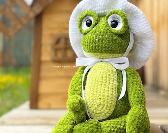 Puede incluir: Una rana verde de peluche hecha a mano, con un sombrero blanco y una cinta. La rana tiene ojos grandes y expresivos y está sentada sobre una superficie de madera. El fondo incluye un molino de viento de madera y vegetación. El texto "PARKAEVA_HM" es visible.