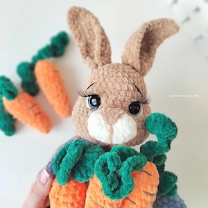 Puede incluir: Un conejo de crochet marr&oacute;n que sostiene tres zanahorias naranjas con hojas verdes. El conejo tiene grandes ojos azules con pesta&ntilde;as largas.