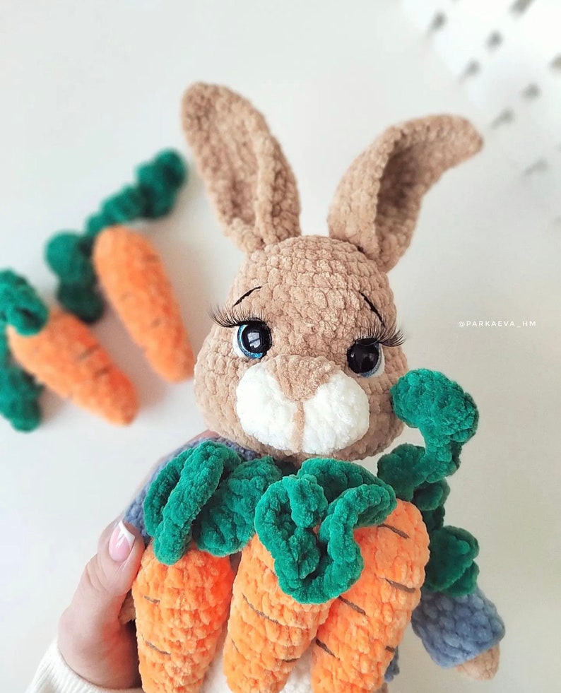Puede incluir: Un conejo de crochet marr&oacute;n que sostiene tres zanahorias naranjas con hojas verdes. El conejo tiene grandes ojos azules con pesta&ntilde;as largas.
