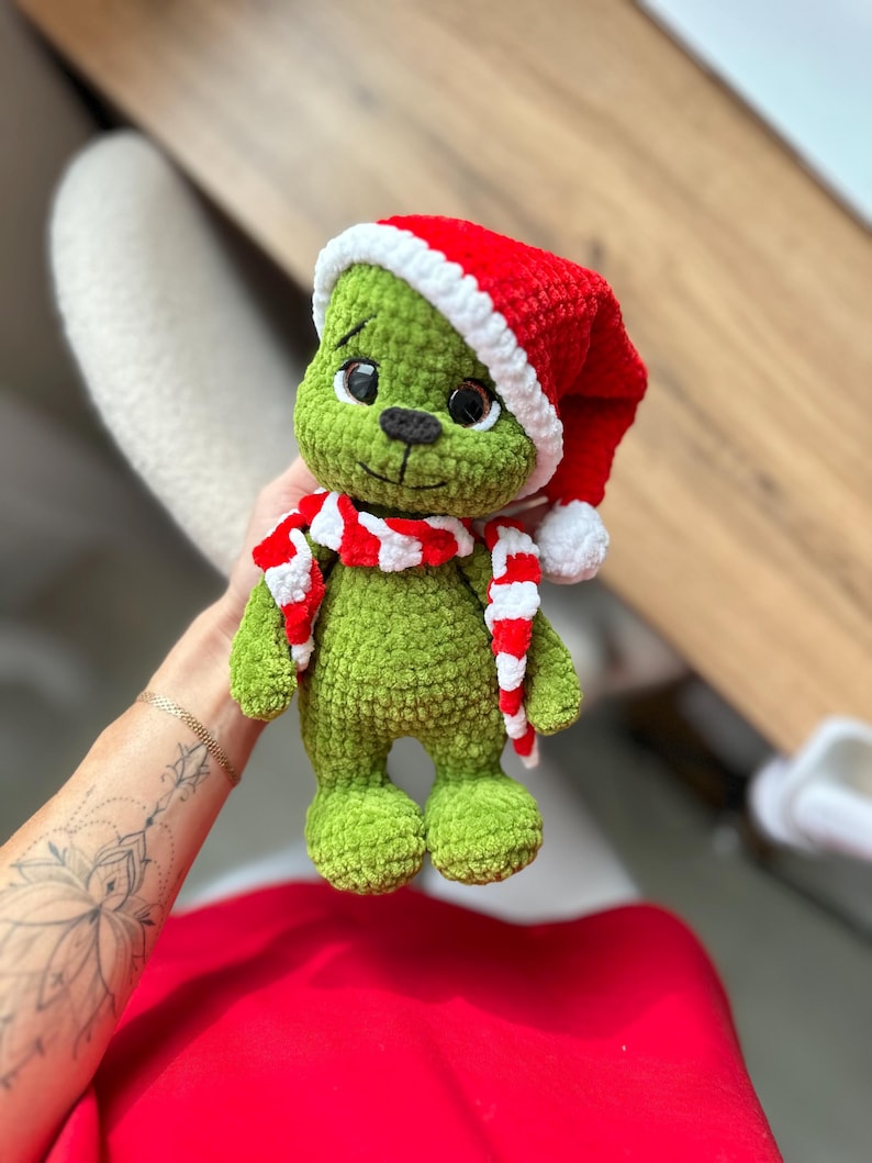 Peut inclure: Une peluche crochet&eacute;e verte avec de grands yeux sombres et un nez noir. La figurine porte un bonnet de P&egrave;re No&euml;l rouge et blanc avec un pompon blanc et une &eacute;charpe ray&eacute;e rouge et blanche assortie. La texture du fil est douce et duveteuse.