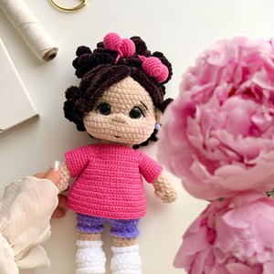 May include: A handmade crochet doll with dark brown hair styled in buns with pink heart-shaped accessories. The doll wears a bright pink short-sleeved shirt, purple shorts, and white shoes. It has large black eyes and a light beige complexion.