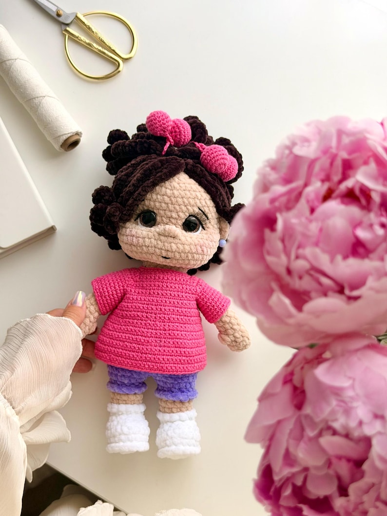 May include: A handmade crochet doll with dark brown hair styled in buns with pink heart-shaped accessories. The doll wears a bright pink short-sleeved shirt, purple shorts, and white shoes. It has large black eyes and a light beige complexion.