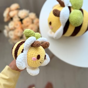 K&ouml;nnte beinhalten: Zwei geh&auml;kelte Bienen-Pl&uuml;schtiere. Die Bienen haben gelbe K&ouml;rper mit braunen Streifen, wei&szlig;e Fl&uuml;gel und gr&uuml;ne Akzente. Eine Biene wird in der Hand gehalten, die andere liegt auf einer wei&szlig;en Oberfl&auml;che. Die Bienen haben l&auml;chelnde Gesichter.