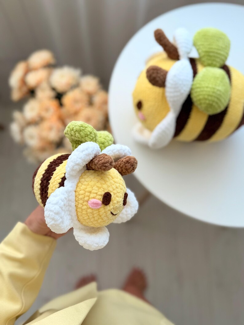 K&ouml;nnte beinhalten: Zwei geh&auml;kelte Bienen-Pl&uuml;schtiere. Die Bienen haben gelbe K&ouml;rper mit braunen Streifen, wei&szlig;e Fl&uuml;gel und gr&uuml;ne Akzente. Eine Biene wird in der Hand gehalten, die andere liegt auf einer wei&szlig;en Oberfl&auml;che. Die Bienen haben l&auml;chelnde Gesichter.