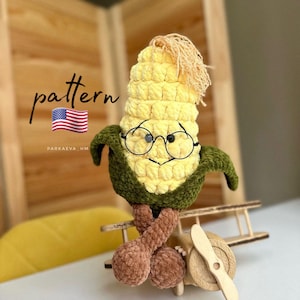May include: A handmade, plush corn character wearing round glasses, with green husks and brown legs. The corn is perched on a wooden airplane. The word "pattern" is written on the image, along with a small American flag.