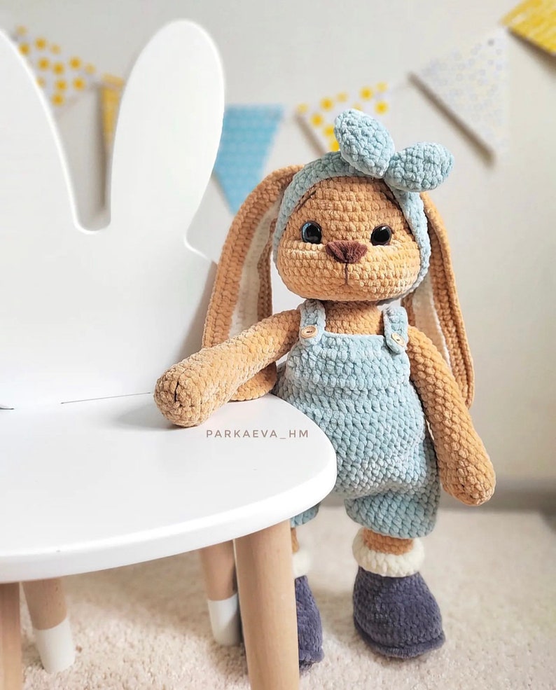 May include: A tan crocheted rabbit plush toy with long ears, wearing a light blue headband with a bow, light blue overalls with small buttons, and dark gray boots with white cuffs. The text PARKAEVA_HM is visible on the rabbit's chest.