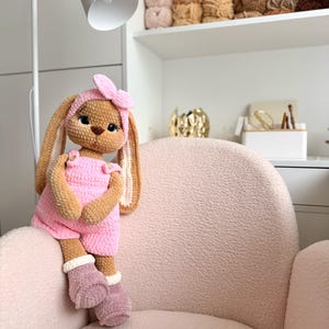 May include: A crocheted stuffed rabbit toy with tan fur, wearing a pink romper with buttons, a matching pink headband with a bow, and mauve-colored boots with white cuffs. The rabbit has large dark eyes, a brown nose, and long, floppy ears.