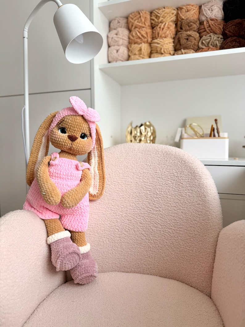 May include: A crocheted stuffed rabbit toy with tan fur, wearing a pink romper with buttons, a matching pink headband with a bow, and mauve-colored boots with white cuffs. The rabbit has large dark eyes, a brown nose, and long, floppy ears.