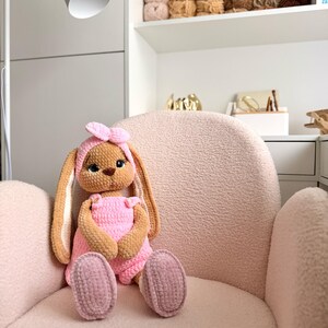 May include: A plush, crocheted rabbit toy with long ears, wearing a pink headband and pink overalls. The rabbit has brown fur, large dark eyes, and a small brown nose. It is crafted from soft, textured yarn and sits upright with its feet forward.
