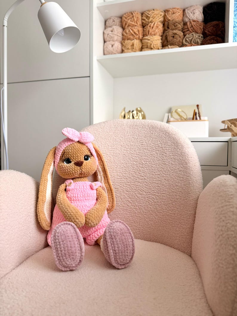 May include: A plush, crocheted rabbit toy with long ears, wearing a pink headband and pink overalls. The rabbit has brown fur, large dark eyes, and a small brown nose. It is crafted from soft, textured yarn and sits upright with its feet forward.