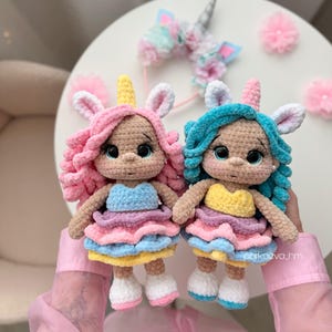 May include: Two handmade crochet unicorn dolls with pink and blue hair, wearing colorful layered dresses. Each doll has a horn, ears, and large eyes. A unicorn headband and pink pom-poms are in the background.