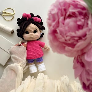 May include: A handmade crochet doll with dark brown curly hair adorned with two pink heart-shaped hair clips. The doll wears a bright pink short-sleeved shirt, purple shorts, and white shoes. It has a light beige complexion with rosy cheeks.