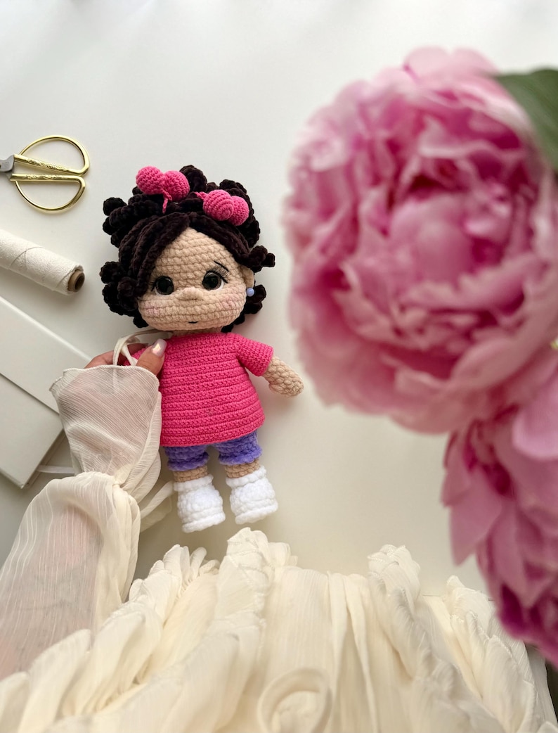 May include: A handmade crochet doll with dark brown curly hair adorned with two pink heart-shaped hair clips. The doll wears a bright pink short-sleeved shirt, purple shorts, and white shoes. It has a light beige complexion with rosy cheeks.