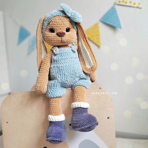 May include: A crocheted plush rabbit toy with tan fur, long ears, and black eyes. The rabbit wears a light blue headband with a bow, light blue overalls with small buttons, and dark purple boots with white cuffs. The text PARKAEVA_HM is visible.