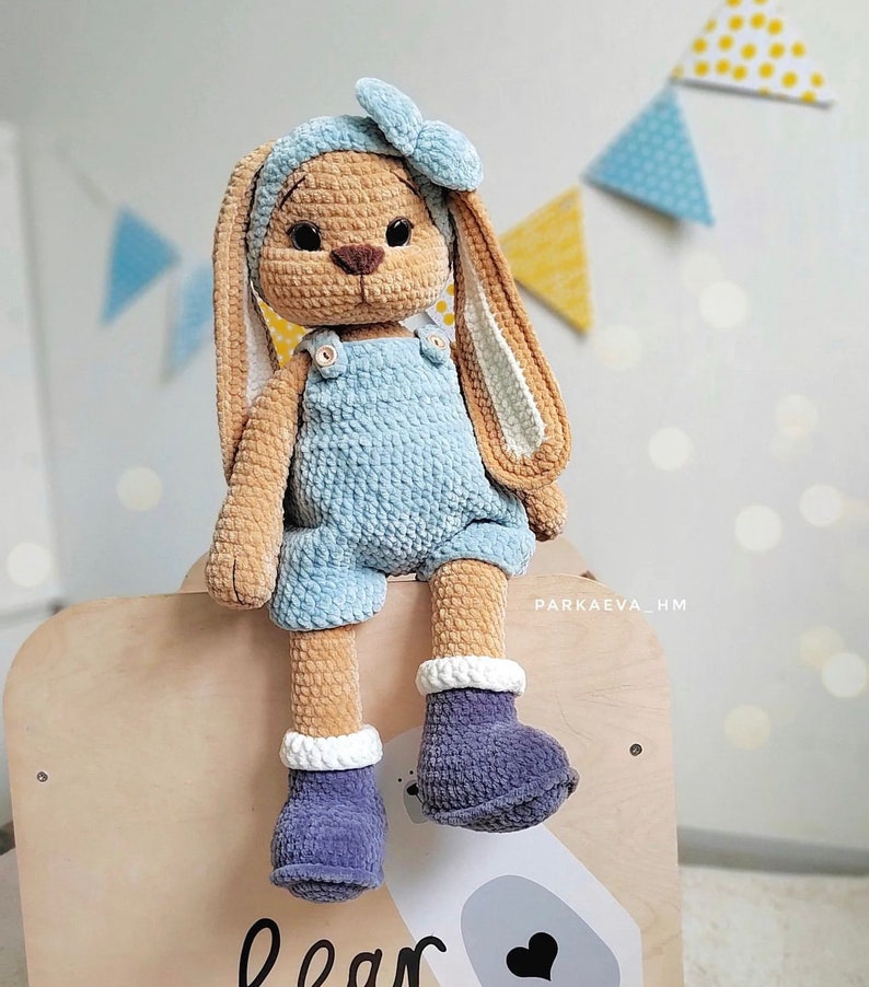 May include: A crocheted plush rabbit toy with tan fur, long ears, and black eyes. The rabbit wears a light blue headband with a bow, light blue overalls with small buttons, and dark purple boots with white cuffs. The text PARKAEVA_HM is visible.