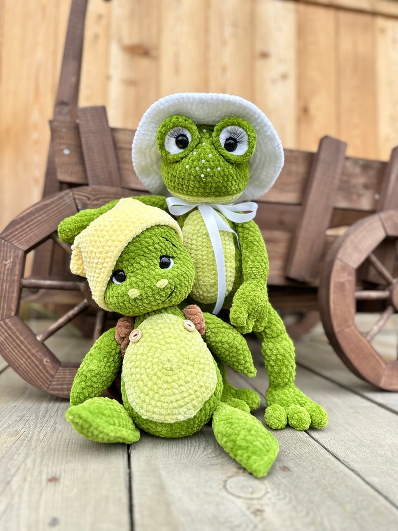 Puede incluir: Dos peluches de rana verdes hechos de hilo suave. La rana m&aacute;s grande lleva un sombrero de ganchillo blanco con una cinta blanca atada bajo la barbilla y tiene pesta&ntilde;as largas. La rana m&aacute;s peque&ntilde;a lleva un gorro de ganchillo amarillo y botones.