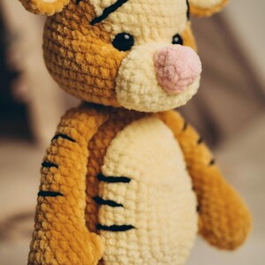 May include: A crocheted tiger plush toy made of soft, textured golden-brown and cream yarn. The stuffed animal features a round head with small ears, black embroidered stripes, a prominent light pink nose, and two small, dark, round plastic eyes.