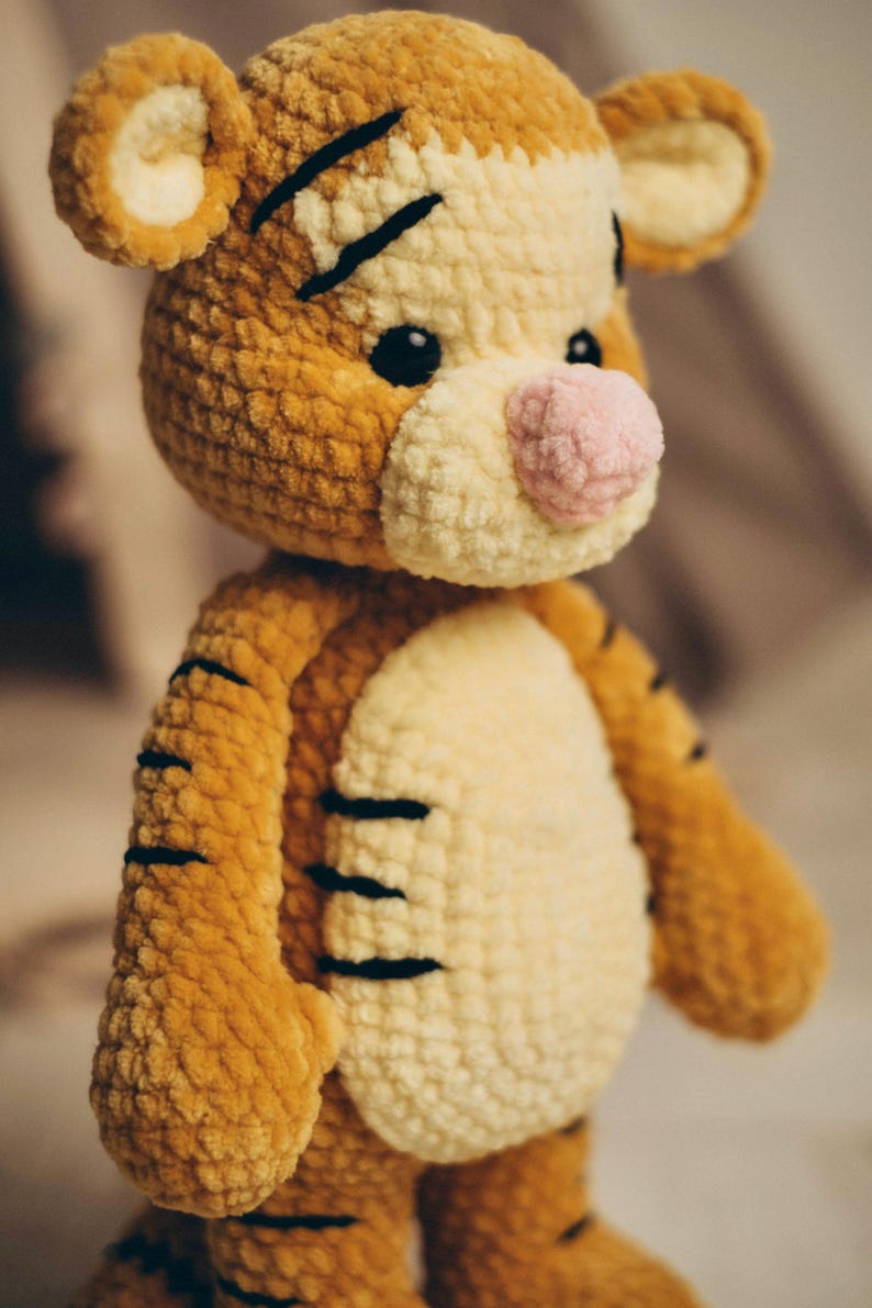 May include: A crocheted tiger plush toy made of soft, textured golden-brown and cream yarn. The stuffed animal features a round head with small ears, black embroidered stripes, a prominent light pink nose, and two small, dark, round plastic eyes.