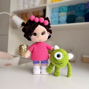 May include: A handmade crochet doll with dark curly hair, a pink headband, a bright pink shirt, purple shorts, and white shoes. The doll stands next to a small, round, green one-eyed monster plush toy with white horns and thin arms and legs.
