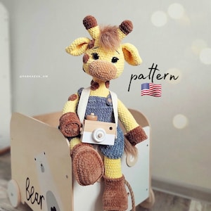 Puede incluir: Un juguete de jirafa de crochet que lleva un mono y sostiene una cámara de juguete. La jirafa está sentada en un vagón de juguete de madera con la palabra "oso" escrita en él. El fondo es una pared clara con la palabra "patrón" escrita en ella.