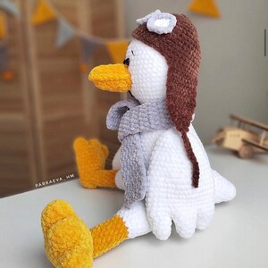 May include: A white crocheted bird wearing a brown crocheted aviator hat with a gray crocheted scarf. The bird has yellow crocheted feet and a yellow crocheted beak.