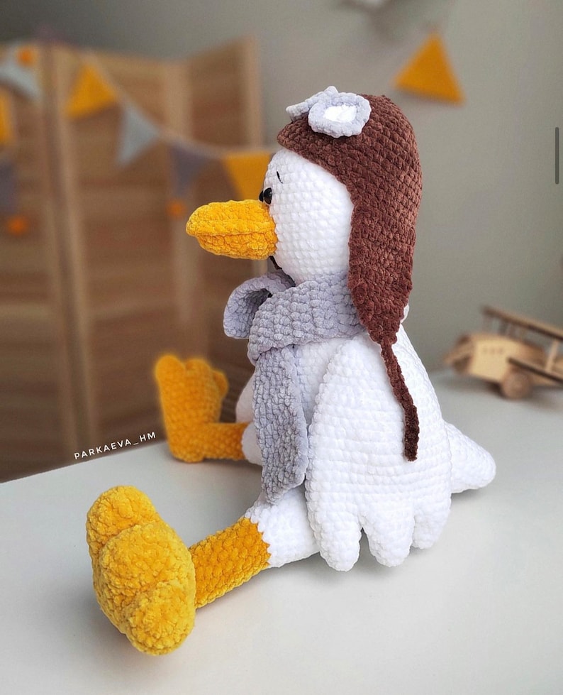 May include: A white crocheted bird wearing a brown crocheted aviator hat with a gray crocheted scarf. The bird has yellow crocheted feet and a yellow crocheted beak.