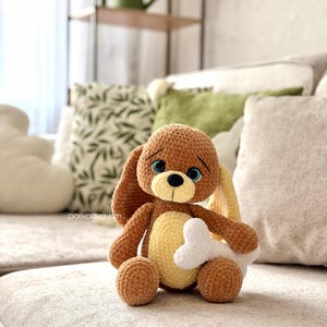 May include: A brown and cream-colored crocheted dog plush toy with large blue eyes, long floppy ears, and a small black nose. The stuffed animal is sitting upright and holding a white crocheted bone in its front paws. The text parkoeva_hm is visible.