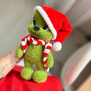 Peut inclure: Une peluche crochet&eacute;e verte &agrave; la texture velout&eacute;e, dot&eacute;e de grands yeux sombres et d'un nez noir. Le jouet porte un bonnet de P&egrave;re No&euml;l rouge vif avec un bord blanc et un pompon blanc, ainsi qu'une &eacute;charpe crochet&eacute;e &agrave; rayures rouges et blanches.