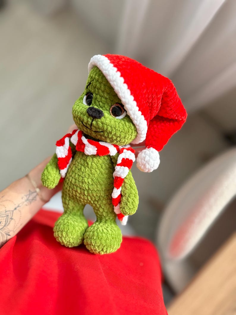 Peut inclure: Une peluche crochet&eacute;e verte &agrave; la texture velout&eacute;e, dot&eacute;e de grands yeux sombres et d'un nez noir. Le jouet porte un bonnet de P&egrave;re No&euml;l rouge vif avec un bord blanc et un pompon blanc, ainsi qu'une &eacute;charpe crochet&eacute;e &agrave; rayures rouges et blanches.