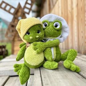 Puede incluir: Dos ranas de peluche hechas de hilo verde texturizado. Una rana lleva un gorro de punto amarillo y tiene la barriga verde claro, mientras que la otra lleva un gorro blanco con cinta, pesta&ntilde;as negras largas y peque&ntilde;os puntos blancos en la cara.