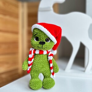 Peut inclure: Une poup&eacute;e en peluche crochet&eacute;e verte &agrave; la texture en relief, dot&eacute;e de grands yeux sombres, d'un nez et d'une bouche brod&eacute;s en noir, portant un bonnet de P&egrave;re No&euml;l rouge et blanc ainsi qu'une &eacute;charpe ray&eacute;e rouge et blanche assortie.