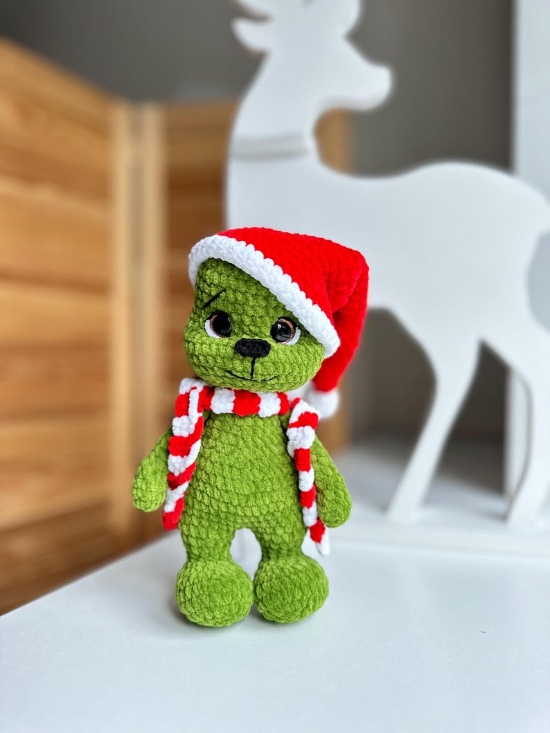 Peut inclure: Une poup&eacute;e en peluche crochet&eacute;e verte &agrave; la texture en relief, dot&eacute;e de grands yeux sombres, d'un nez et d'une bouche brod&eacute;s en noir, portant un bonnet de P&egrave;re No&euml;l rouge et blanc ainsi qu'une &eacute;charpe ray&eacute;e rouge et blanche assortie.