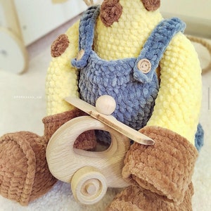 May include: A crocheted giraffe toy wearing blue overalls and holding a wooden toy airplane. The giraffe has brown spots and a yellow body. The airplane has a brown propeller and a wooden wheel.