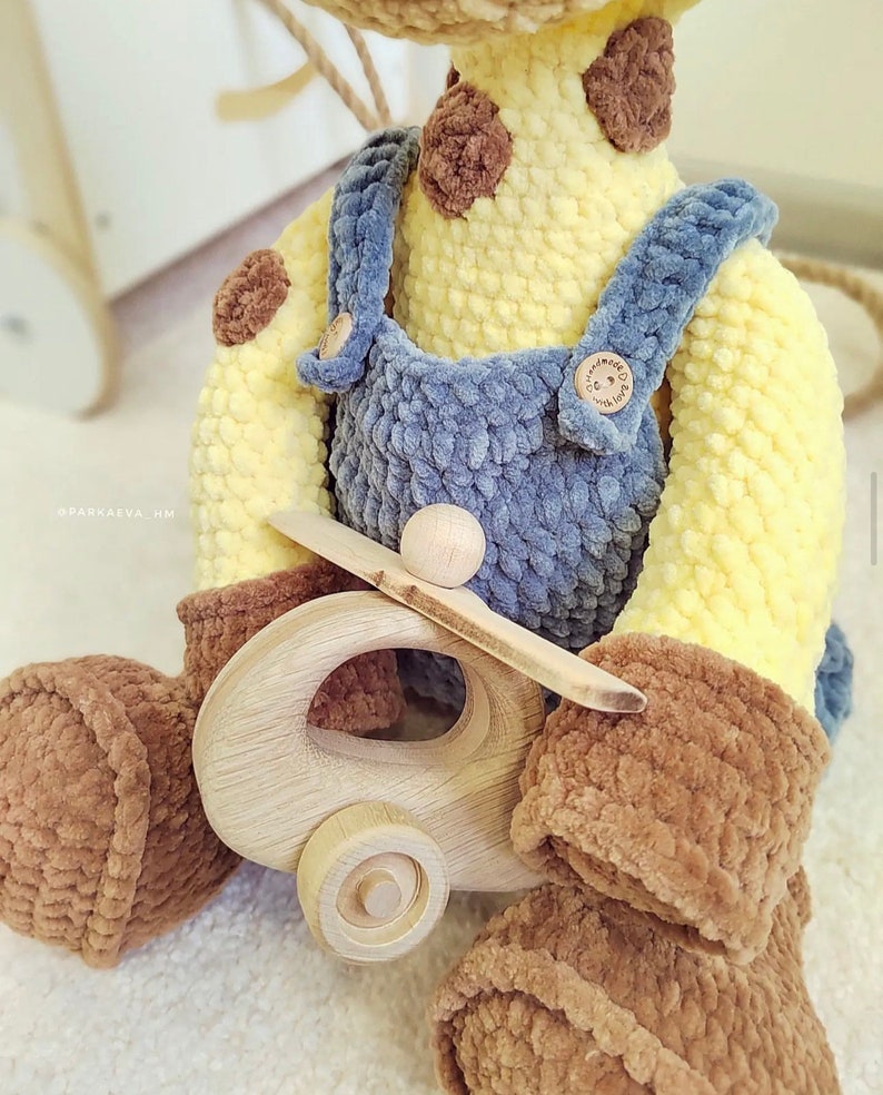 May include: A crocheted giraffe toy wearing blue overalls and holding a wooden toy airplane. The giraffe has brown spots and a yellow body. The airplane has a brown propeller and a wooden wheel.