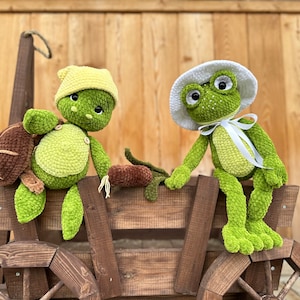 Puede incluir: Un peluche de tortuga de ganchillo con gorro amarillo y un peluche de rana de ganchillo con sombrero blanco y cinta blanca, ambos sentados en un carro de madera. La tortuga tiene un caparaz&oacute;n marr&oacute;n y la rana tiene ojos grandes.