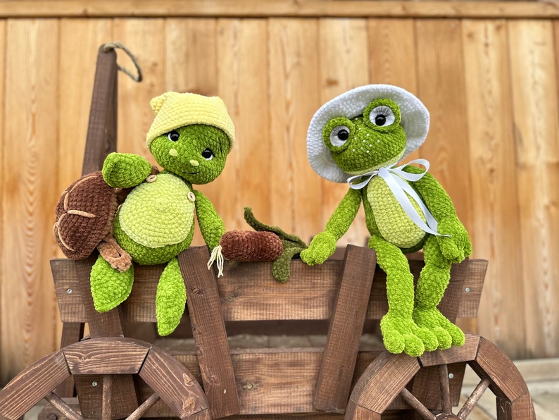 Puede incluir: Un peluche de tortuga de ganchillo con gorro amarillo y un peluche de rana de ganchillo con sombrero blanco y cinta blanca, ambos sentados en un carro de madera. La tortuga tiene un caparaz&oacute;n marr&oacute;n y la rana tiene ojos grandes.