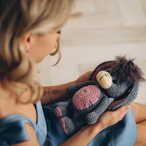 Pu&ograve; includere: Una donna con un vestito blu tiene in mano un peluche di colore grigio e rosa lavorato a uncinetto con pelliccia marrone. L'animale ha grandi orecchie e un lungo muso.