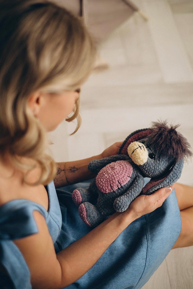 Pu&ograve; includere: Una donna con un vestito blu tiene in mano un peluche di colore grigio e rosa lavorato a uncinetto con pelliccia marrone. L'animale ha grandi orecchie e un lungo muso.