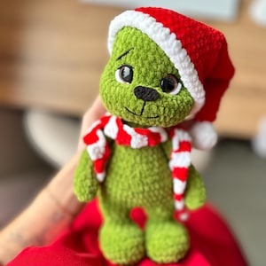 Peut inclure: Une poup&eacute;e en peluche crochet&eacute;e verte &agrave; la texture en relief, dot&eacute;e de grands yeux sombres, d'un nez noir et d'une bouche brod&eacute;e. La poup&eacute;e porte un bonnet de P&egrave;re No&euml;l crochet&eacute; rouge et blanc avec un pompon blanc et une &eacute;charpe ray&eacute;e assortie.