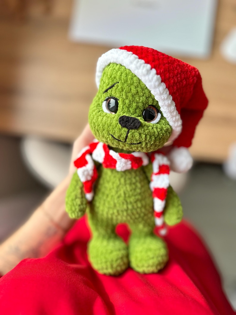 Peut inclure: Une poup&eacute;e en peluche crochet&eacute;e verte &agrave; la texture en relief, dot&eacute;e de grands yeux sombres, d'un nez noir et d'une bouche brod&eacute;e. La poup&eacute;e porte un bonnet de P&egrave;re No&euml;l crochet&eacute; rouge et blanc avec un pompon blanc et une &eacute;charpe ray&eacute;e assortie.