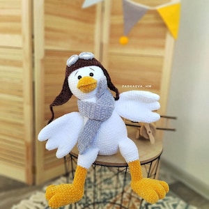 May include: A white crocheted goose wearing a brown aviator hat with goggles, a gray scarf, and yellow feet. The goose is sitting on a black metal stool with a woven seat.
