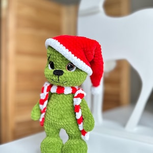 Peut inclure: Une poup&eacute;e en peluche crochet&eacute;e verte avec de grands yeux marron et un nez cousu noir. La poup&eacute;e porte un bonnet de P&egrave;re No&euml;l crochet&eacute; rouge et blanc ainsi qu'une &eacute;charpe assortie ray&eacute;e rouge et blanche. La texture du fil est douce et duveteuse.