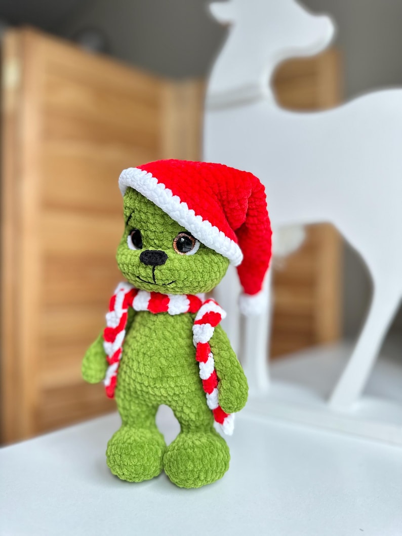 Peut inclure: Une poup&eacute;e en peluche crochet&eacute;e verte avec de grands yeux marron et un nez cousu noir. La poup&eacute;e porte un bonnet de P&egrave;re No&euml;l crochet&eacute; rouge et blanc ainsi qu'une &eacute;charpe assortie ray&eacute;e rouge et blanche. La texture du fil est douce et duveteuse.